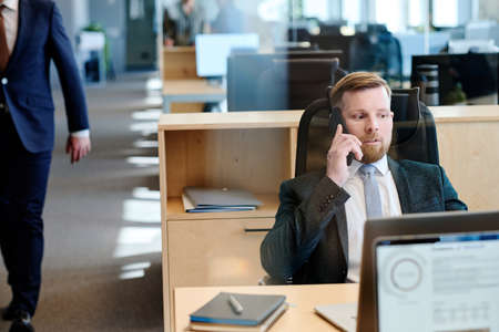 Young bearded manager in suit having a conversation on mobile phone while working on laptop at...