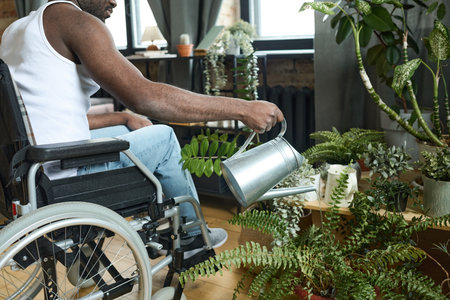 Close-up of African young man with disability sitting in wheelchair and watering house plants from watering can in the roomの写真素材