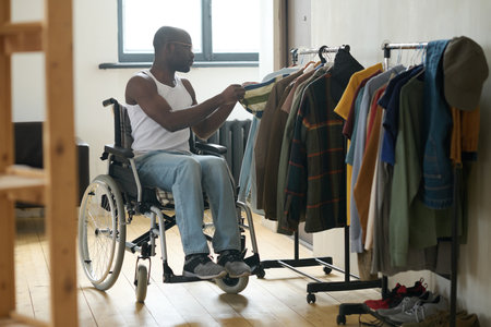 African man with disability sitting in wheelchair and choosing clothes to wear hanging on rackの写真素材