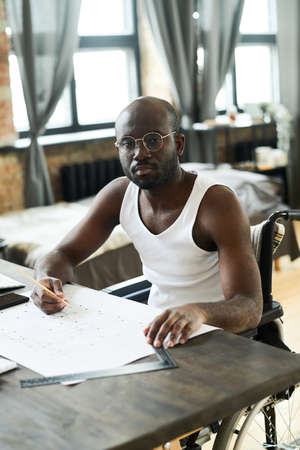 Portrait of African young architect with disability looking at camera while sitting at table and...