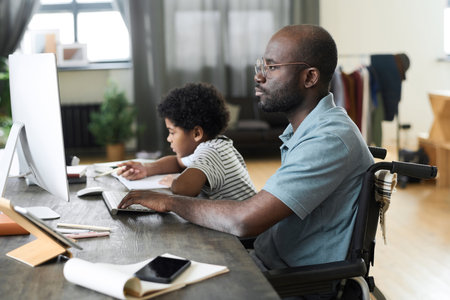 African disabled father working online on computer sitting in wheelchair at desk with his son doing homework in backgroundの写真素材