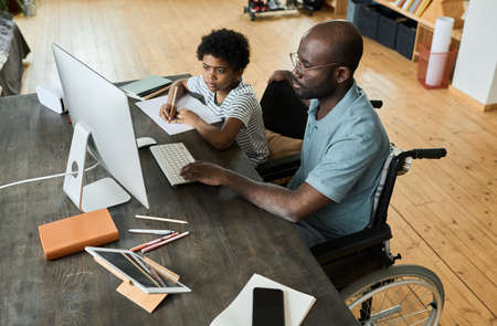 African disabled father sitting in wheelchair using computer at table to help his son with homeworkの写真素材