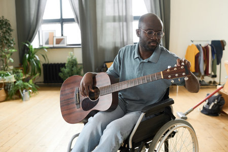 African American man with disability sitting in wheelchair and tuning a guitar for playing music at...