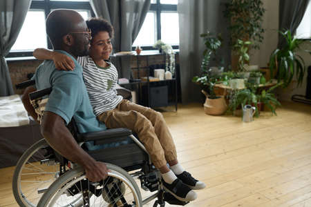 African disabled father riding his son on wheelchair while he sitting on his knees, they playing in the roomの写真素材