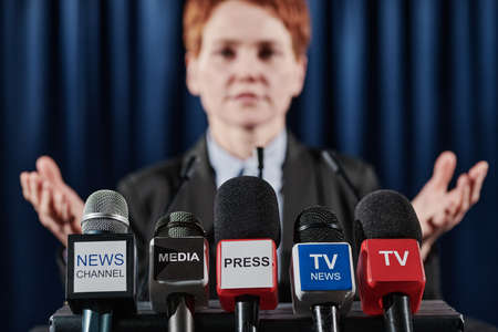 Close-up of microphones of various television media with speaker standing behind them at tribuneの写真素材