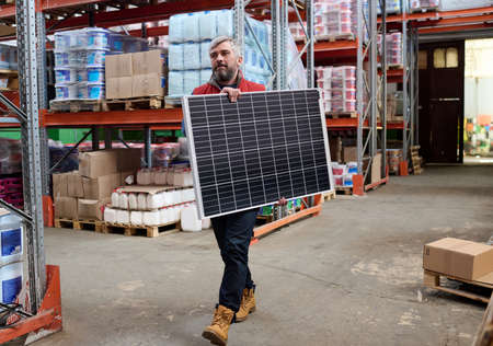 Mature bearded loader walking along the shelves with parcels with a solar battery in his hands in warehouseの写真素材