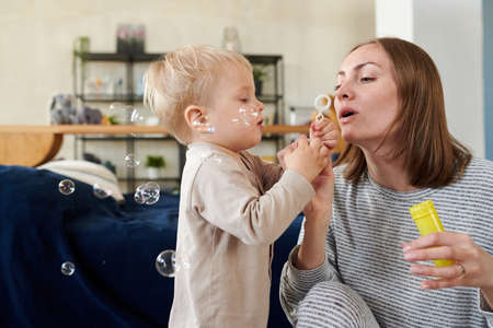 Young mother blowing soap bubbles while her little son trying to catch them during their games at homeの写真素材