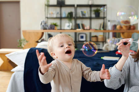 Adorable little kid catching soap bubbles blowing by his mom in the roomの写真素材