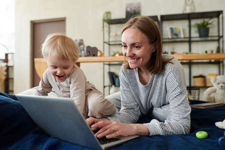 Smiling young mother typing on laptop and doing her online work while her child playing next to her...