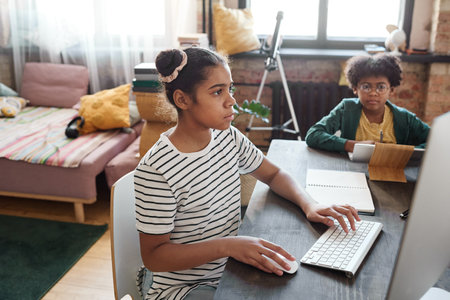 African girl using computer while sitting at table together with her brother during distance...