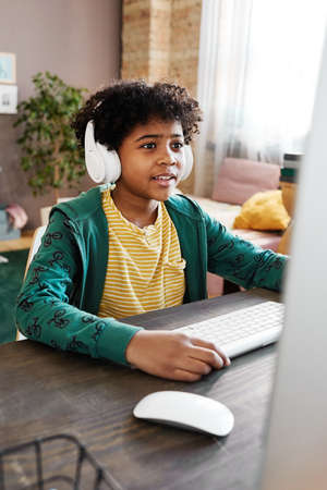 Smiling African boy in wireless headphones sitting in front of computer monitor and playing video...