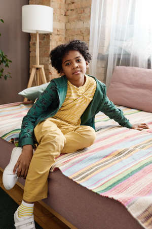Portrait of African little boy in casual clothing sitting on sofa and looking at camera during his leisure timeの写真素材