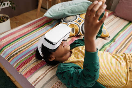 African boy lying on sofa in VR goggles playing virtual reality game and gesturingの写真素材