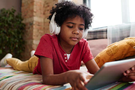 African little boy in wireless headphones lying on sofa and watching video online using digital tabletの写真素材