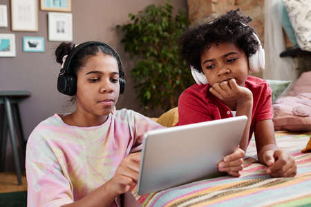 Brother and sister in wireless headphones watching video online on digital tablet together at home