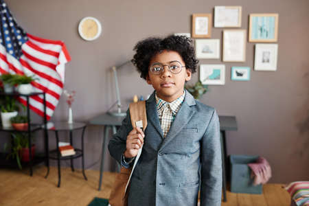 Portrait of African little schoolboy in suit looking at camera holding school bag behind his back standing in the roomの写真素材
