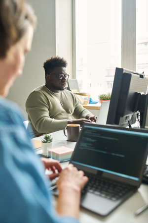 African young man sitting at table in front of computer monitor and typing with his colleague...