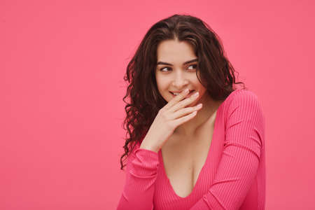 Portrait of funny young woman closed her mouth with hand while smiling against the pink background