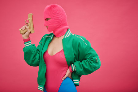 Young criminal girl in pink balaclava and body posing with weapon against pink backgroundの写真素材