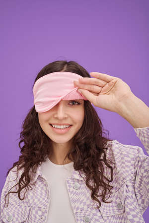 Portrait of young girl with wavy hair peeping from sleep mask and smiling at camera on purple backgroundの写真素材