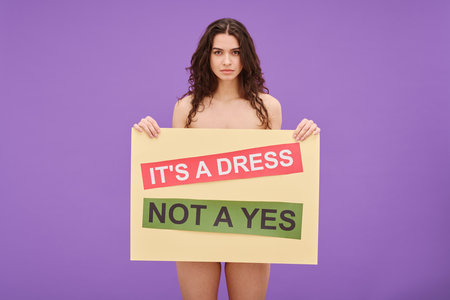 Portrait of young shirtless woman holding Its a dress not a yes poster in front of her standing against color backgroundの写真素材