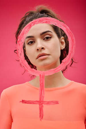 Portrait of young girl looking at camera through venus sign drawed by lipstick against pink backgroundの写真素材