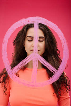 Portrait of young peaceful woman with closed eyes standing in front of pacifism sign on pink backgroundの写真素材