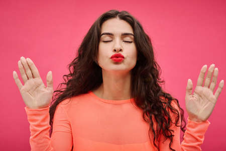 Portrait of young woman with red lipstick leaning against the transparent wall and kissing on pink backgroundの写真素材
