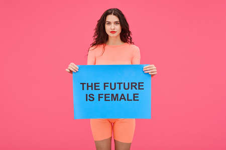 Portrait of young woman holding The future is female poster and looking at camera expressing her rights against pink backgroundの写真素材