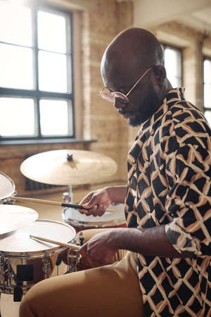 African man in eyeglasses using drumsticks to play drums during practice at studioの写真素材