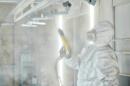 Worker in protective suit working with spray gun to paint metal details at factoryの写真素材