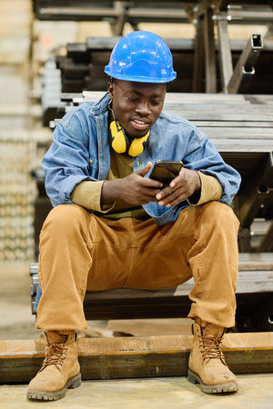 African young worker in hardhat sitting on metal sticks watching video in smartphone during his...