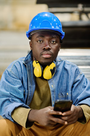Portrait of African young male worker in protective hardhat looking at camera while playing on his...