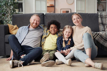 Portrait of happy foster parents sitting on floor with their adopted children and smiling at camera, they resting at homeの写真素材