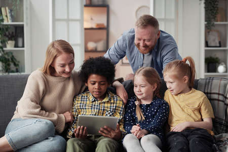 Happy parents spending time with their adopted children using digital tablet together on sofa in living roomの写真素材