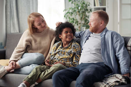 Young parents happy to adopt little boy, they sitting on sofa and talking to each other during their...