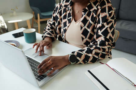 Close-up of African girl doing her online work typing on laptop while sitting at table with notepad and cup of coffee at homeの写真素材