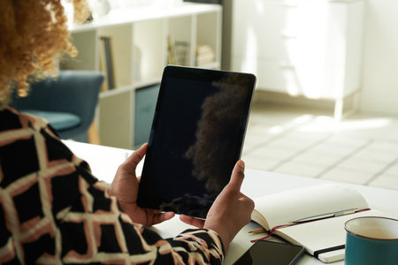 Close-up of African girl looking at screen of digital tablet and working online sitting at table...