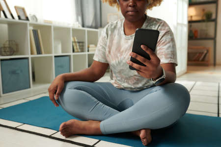 Close-up of African young woman sitting on exercise mat and watching sports training online using...