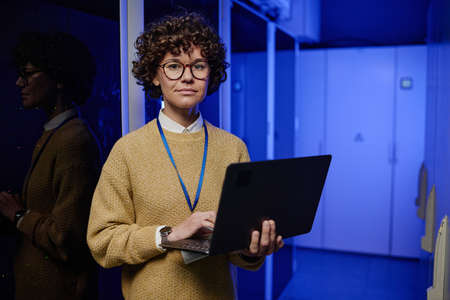 Portrait of female technician in eyeglasses using her laptop while working in data center with...