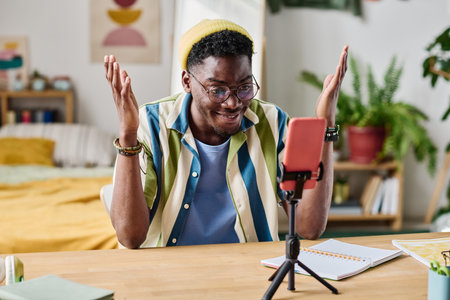 African excited blogger speaking looking at smartphone on tripod at home table while recording blogの写真素材