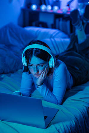 Teenage girl in headphones lying on her bed and playing computer game at night in her bedroomの写真素材