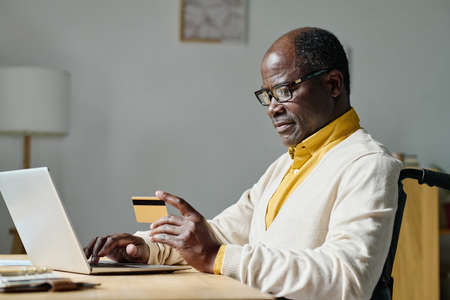 African man in eyeglasses paying online with credit card using laptop at table sitting in wheelchair