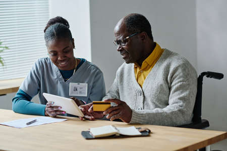 African senior man learning to pay online with credit card using digital tablet together with...