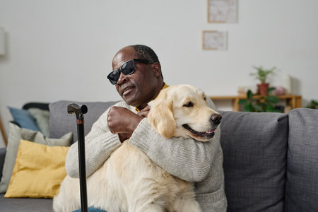 African senior man in glasses with bad sight embracing his individual guide dog while sitting on sofa at homeの写真素材