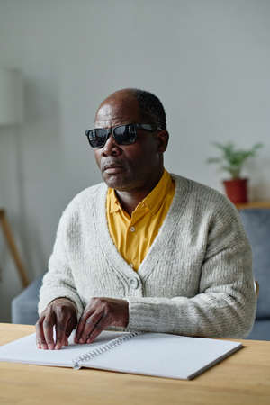 African blind man in dark glasses sitting at table and reading book in braille languageの写真素材
