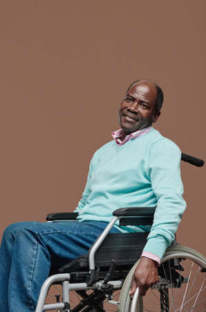 Portrait of African senior man with disability sitting in wheelchair against beige background