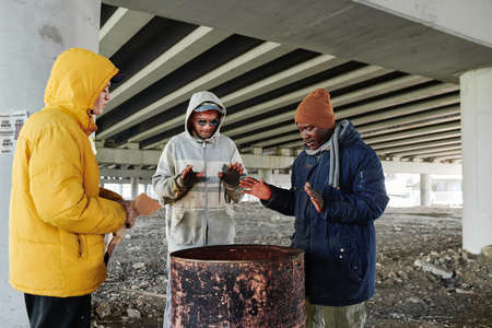 Group of beggars in dirty clothing standing near the barrel with faire and warming together in cold day outdoorsの写真素材