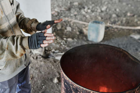 Close-up of poor man in torn gloves and dirty clothes warming up by fire standing near the barrel in the cityの写真素材