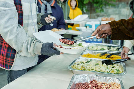 Close-up of volunteers serving homeless people and putting meal on plates, they feeding poor people during charityの写真素材